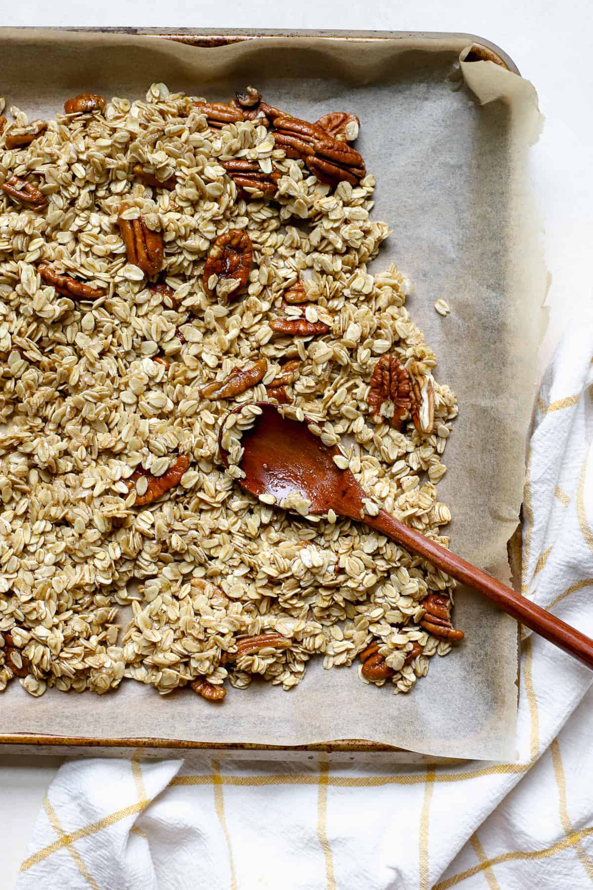 Fully baked maple pecan granola on sheet pan lined with unbleached parchment paper, and wooden spoon resting in pan, set on golden yellow and white window-pane linen.