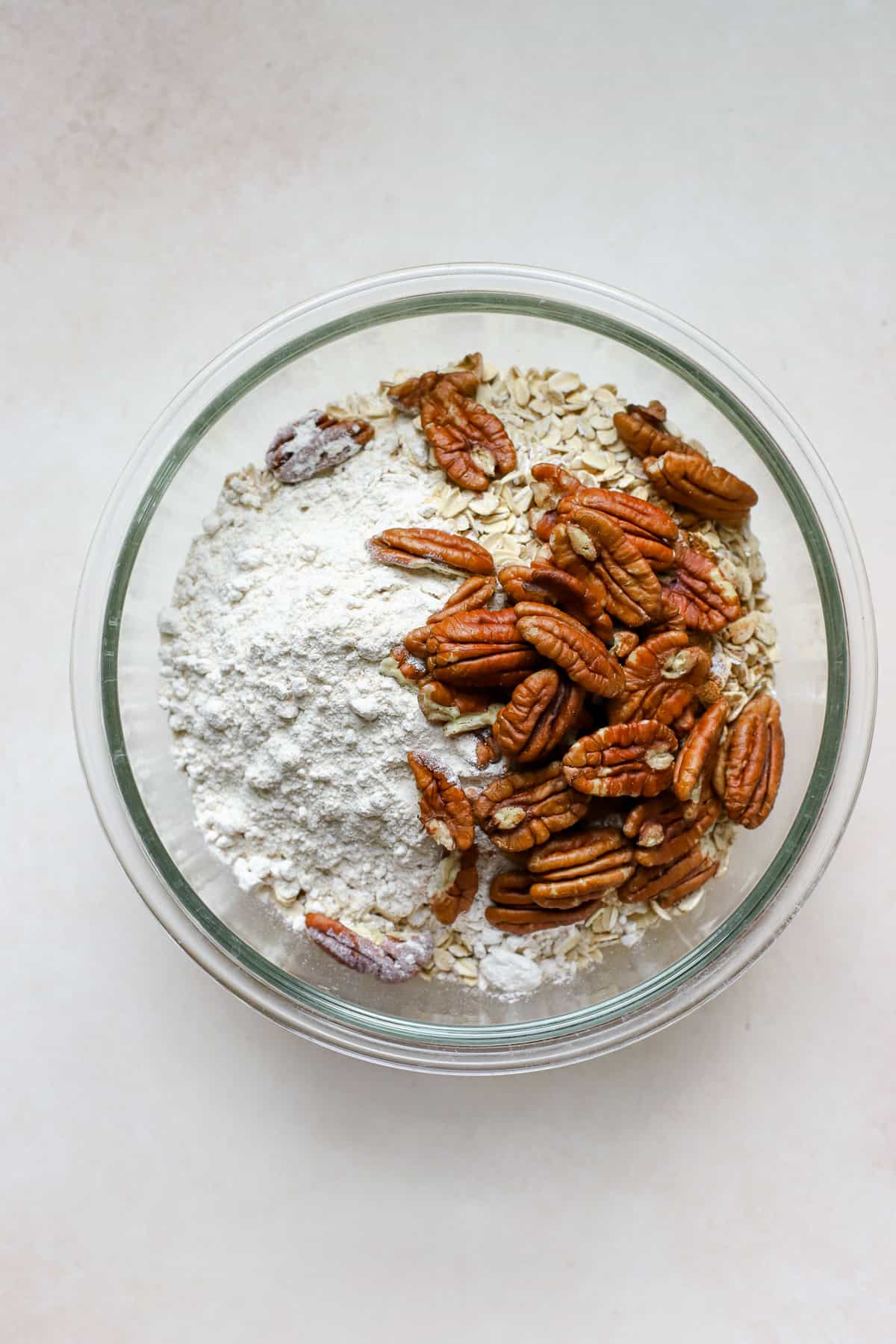 Maple pecan granola dry ingredients in clear glass bowl, including oats, flour, cinnamon, salt, and pecans, on beige surface.