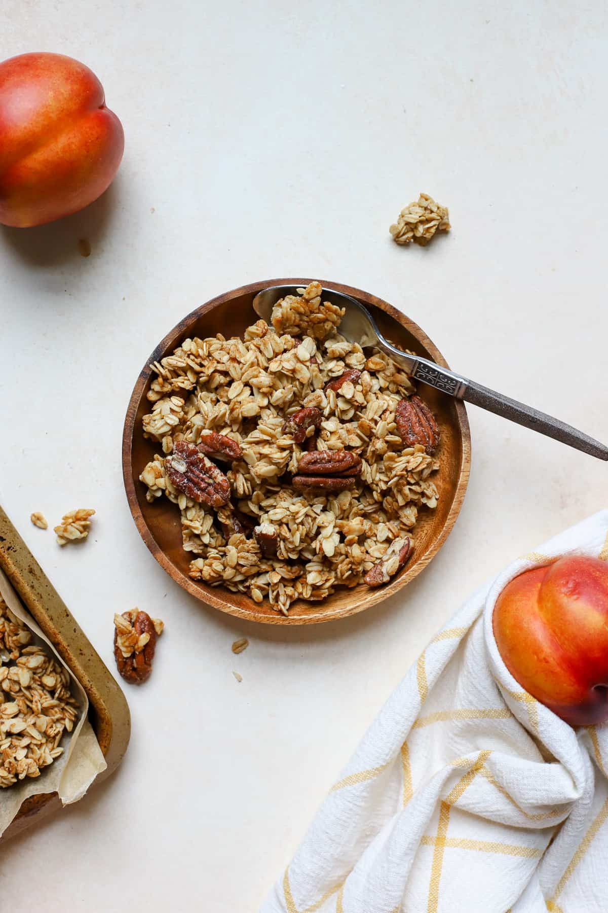 Maple pecan granola on small wooden plate with spoon and two whole peaches next to it, corner of sheet pan of granola peaking in from the left side, on beige surface and golden yellow and white linen.