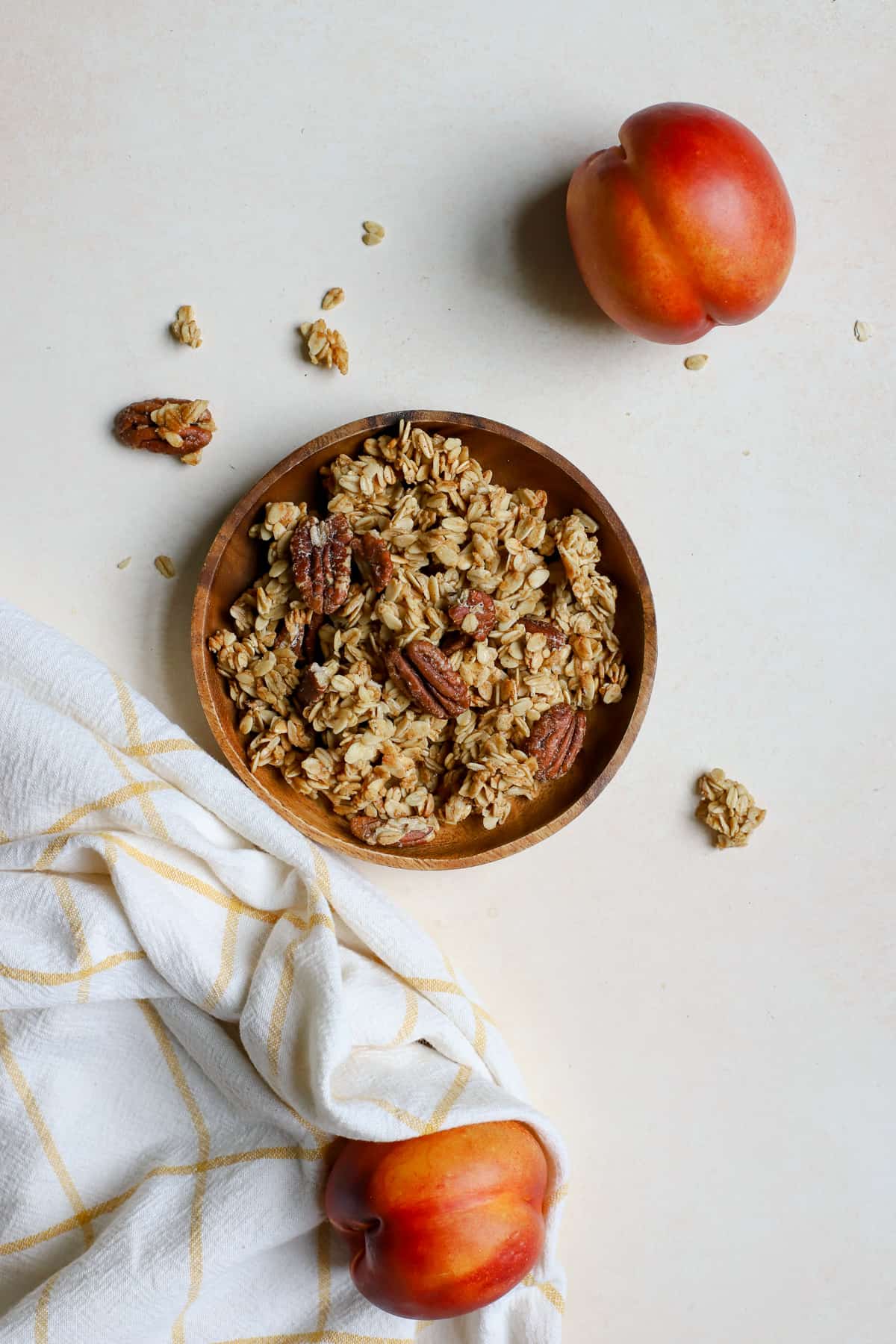 Maple pecan granola on small wooden plate with two whole peaches next to it, on beige surface and golden yellow and white linen.