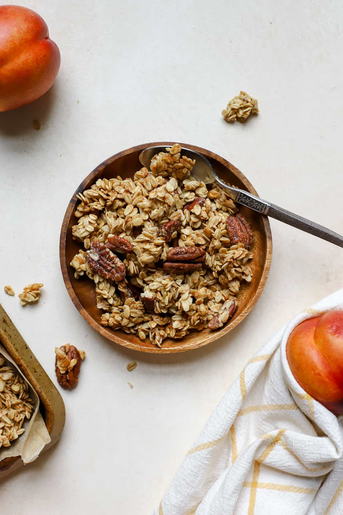 Maple pecan granola on small wooden plate with spoon and two whole peaches next to it, corner of sheet pan of granola peaking in from the left side, on beige surface and golden yellow and white linen.