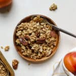 Maple pecan granola on small wooden plate with spoon and two whole peaches next to it, corner of sheet pan of granola peaking in from the left side, on beige surface and golden yellow and white linen.