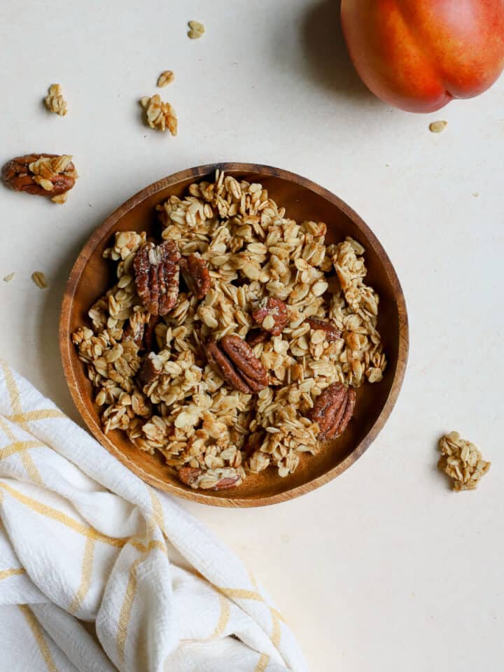 Maple pecan granola on small wooden plate with spoon and two whole peaches next to it, corner of sheet pan of granola peaking in from the left side, on beige surface and golden yellow and white linen.