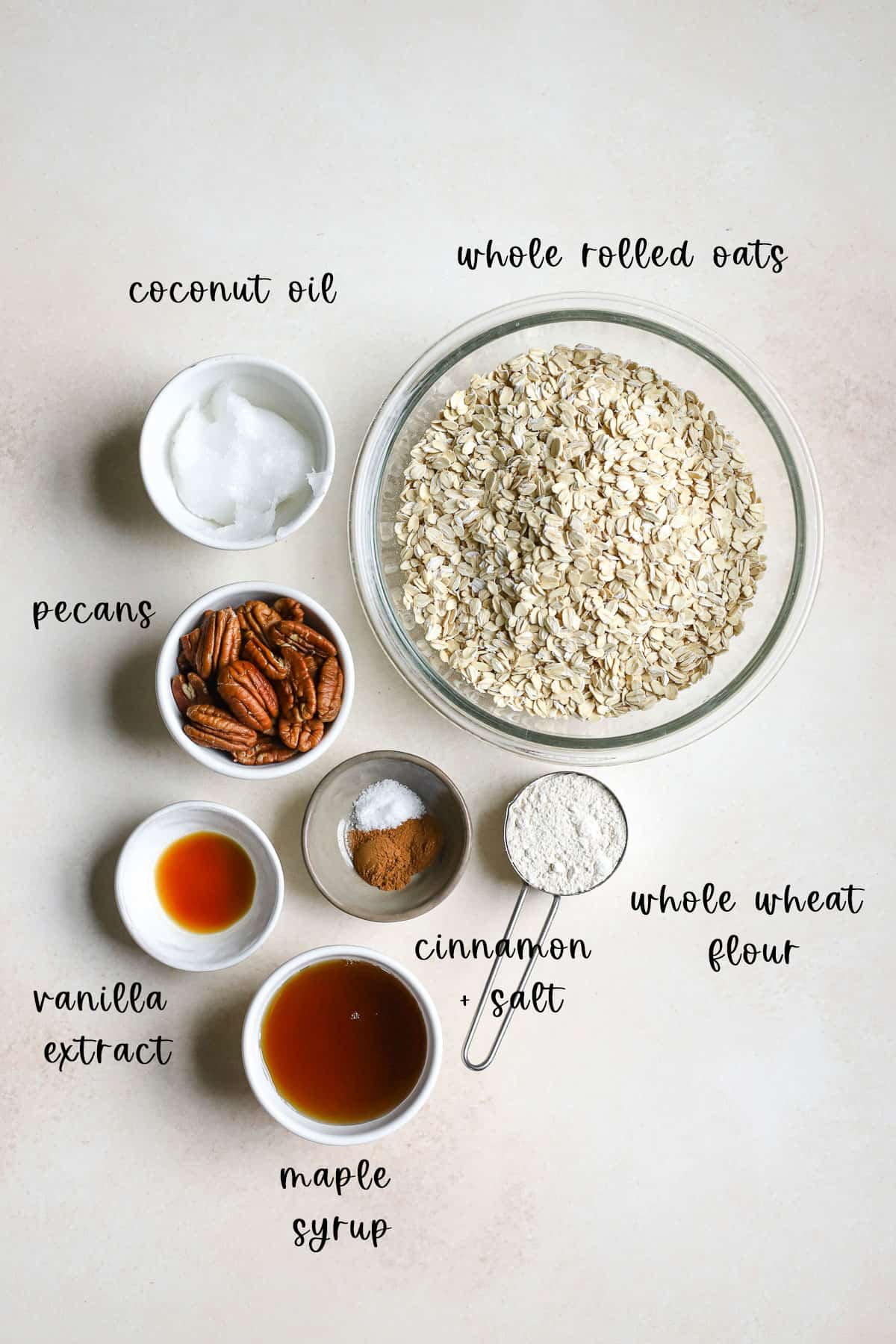 Maple pecan granola ingredients individually measured out including oats, coconut oil, whole wheat flour, cinnamon, salt, vanilla extract, maple syrup, and pecans, on beige surface.