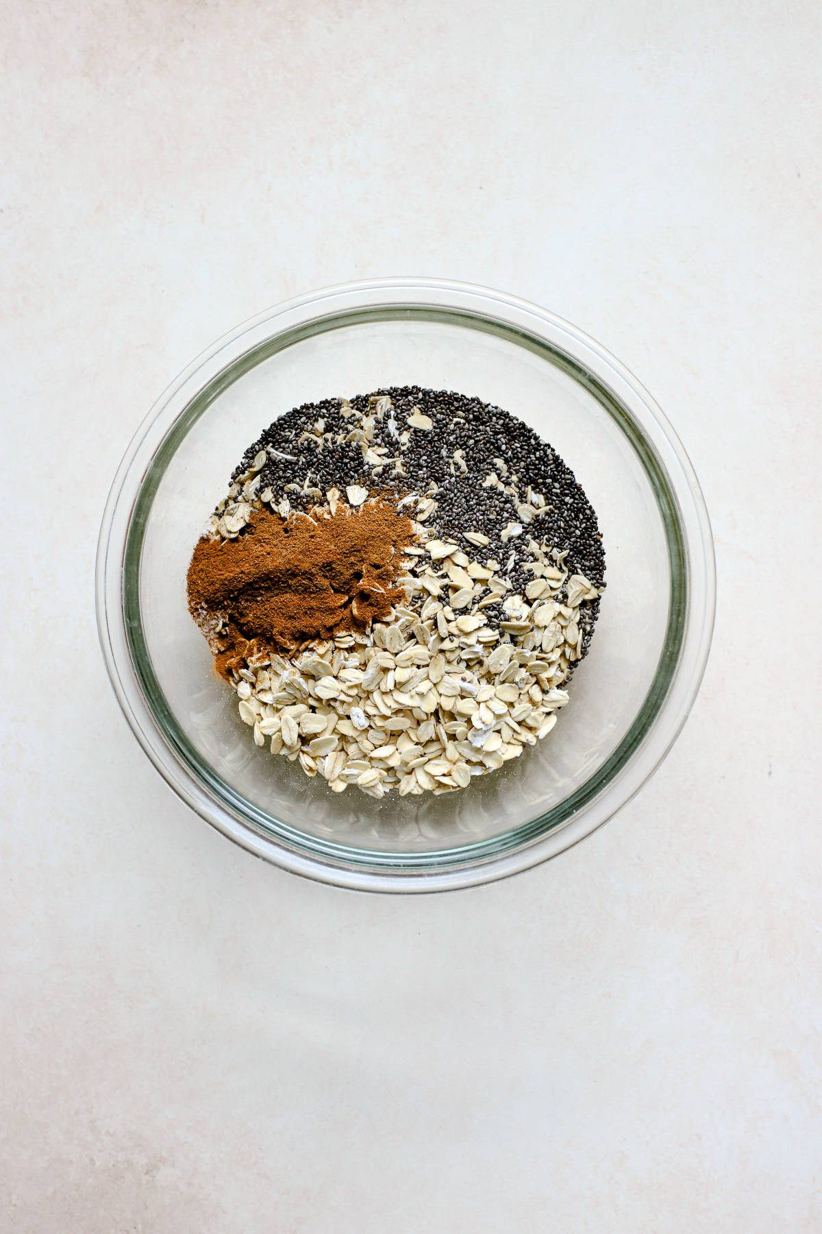 Whole rolled oats, chia seeds, pumpkin pie spice, cinnamon, and salt in a clear glass bowl on beige and white surface.