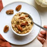 Pumpkin pie overnight oats topped with pecans in a small white bowl sprinkled with cinnamon, and hand holding spoon scooping into oats.