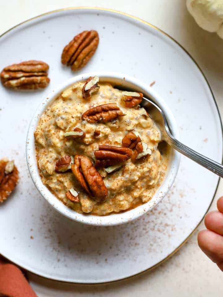 Pumpkin pie overnight oats topped with pecans in a small white bowl sprinkled with cinnamon, and hand holding spoon scooping into oats.