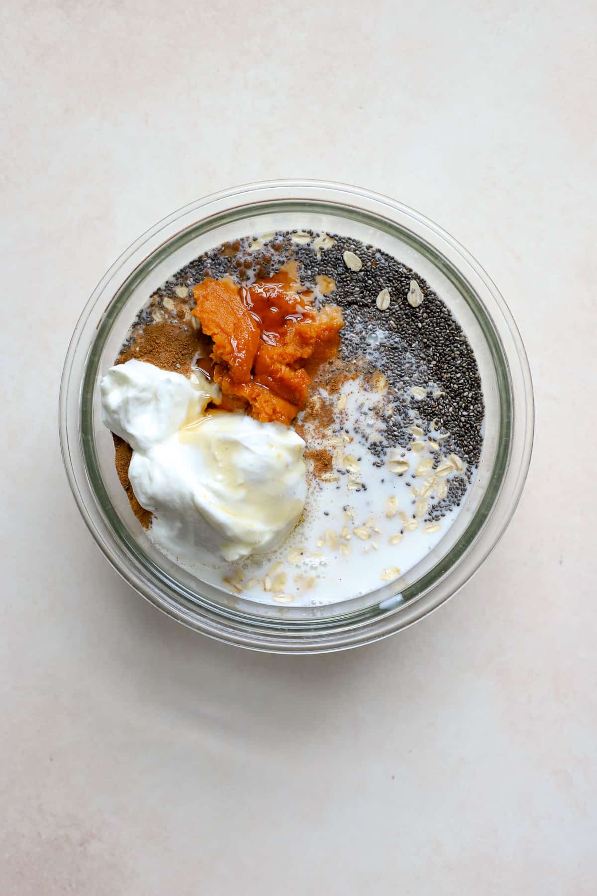 Pumpkin overnight oats ingredients added to a clear glass bowl before being mixed together, on beige and white surface.