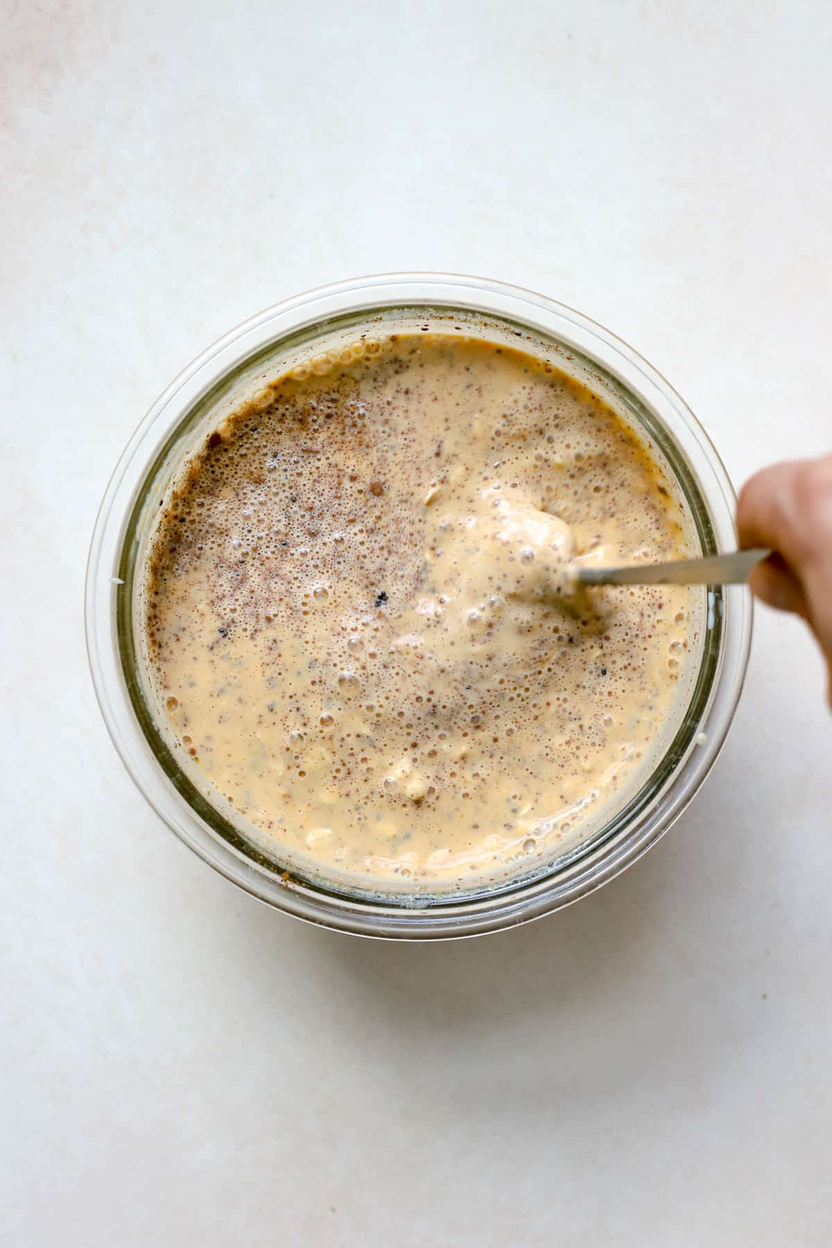 All overnight oats ingredients mixed together in a clear glass bowl being stirred with a spoon, on a beige and white surface.