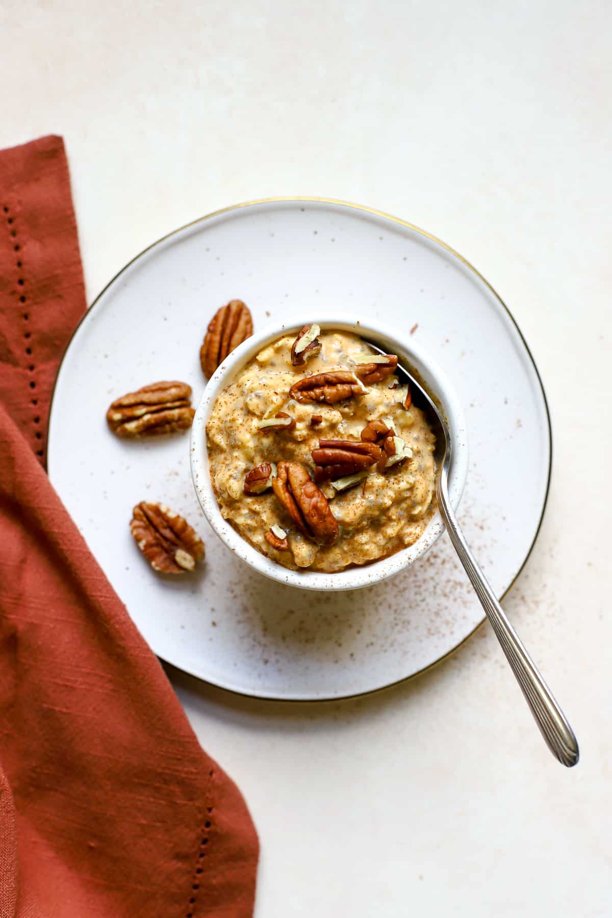 Pecan-topped pumpkin pie overnight oats sprinkled with cinnamon in a small white bowl with spoon, on white, gold-flecked plate with rust-colored linen on the side.