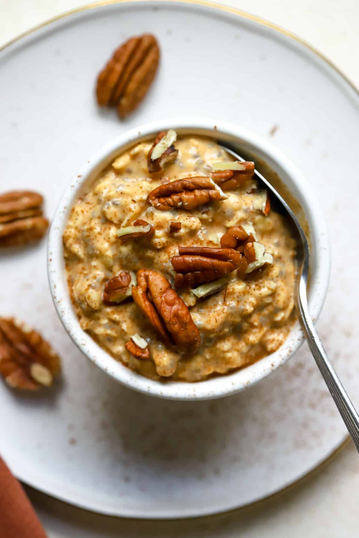 Pecan-topped pumpkin pie overnight oats sprinkled with cinnamon in a small white bowl with spoon, on white, gold-flecked plate.