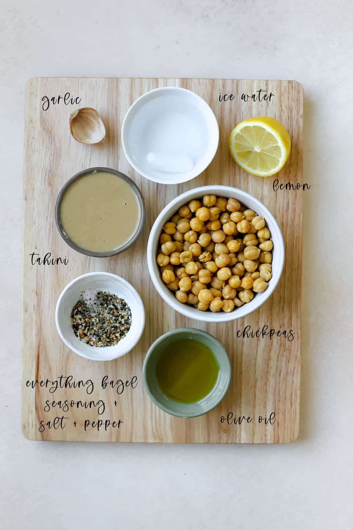 Hummus ingredients on wooden cutting board, including chickpeas, tahini, a clove of garlic, half a lemon, iced water, olive oil, everything bagel seasoning, salt, and pepper. Everything is measured out and prepared to make hummus recipe.