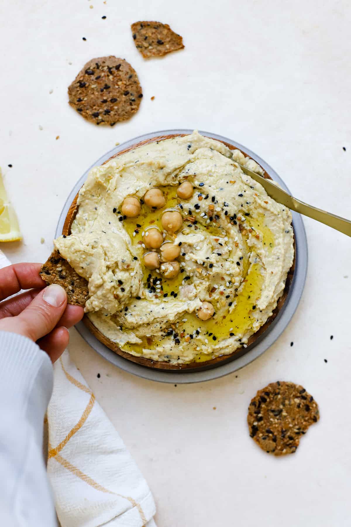 Everything bagel hummus served on wooden plate, drizzled with olive oil, with round seedy crackers on the side and being dipped.