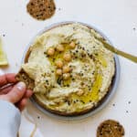 Everything bagel hummus served on wooden plate, drizzled with olive oil, with round seedy crackers on the side and being dipped.