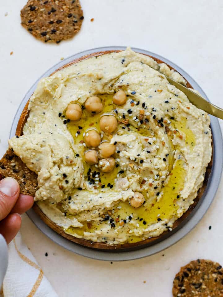 Everything bagel hummus served on wooden plate, drizzled with olive oil, with round seedy crackers on the side and being dipped.