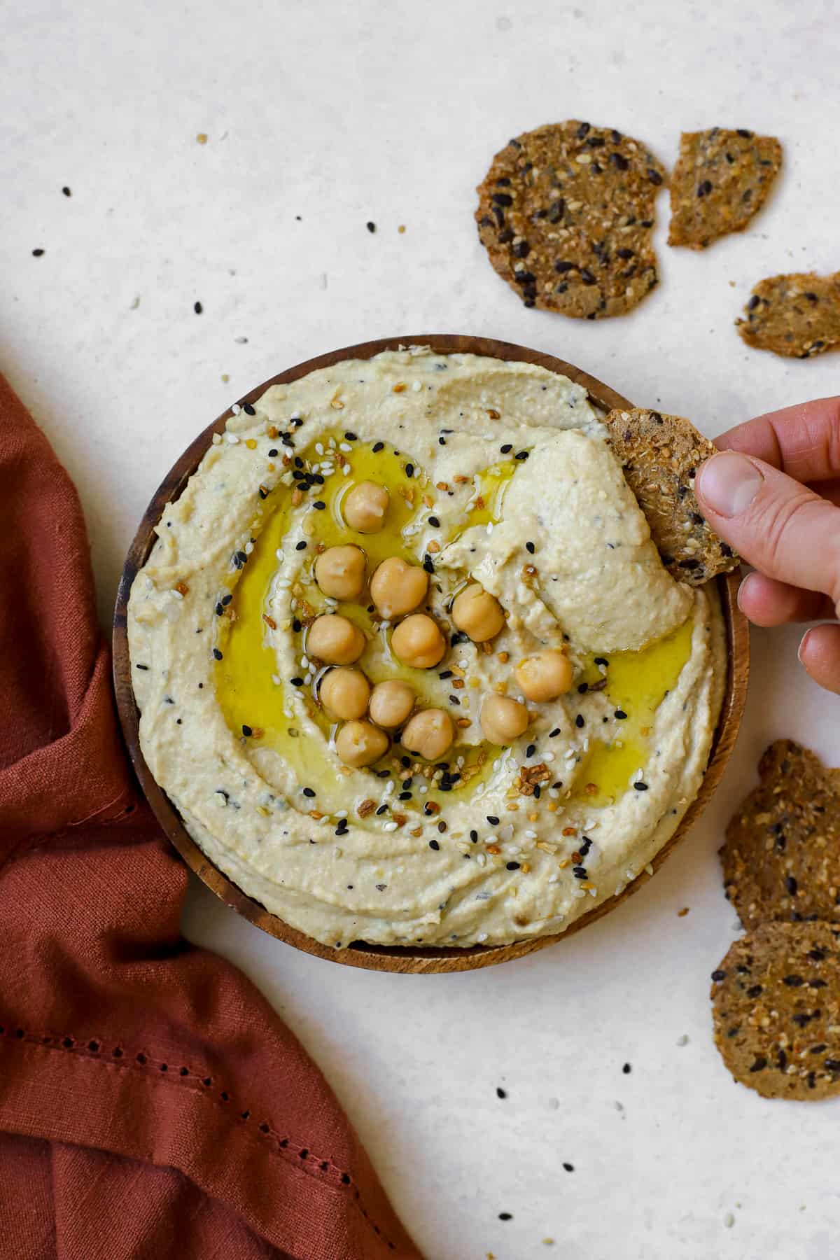 Everything bagel hummus served on wooden plate, drizzled with olive oil, with round seedy crackers on the side and being dipped.