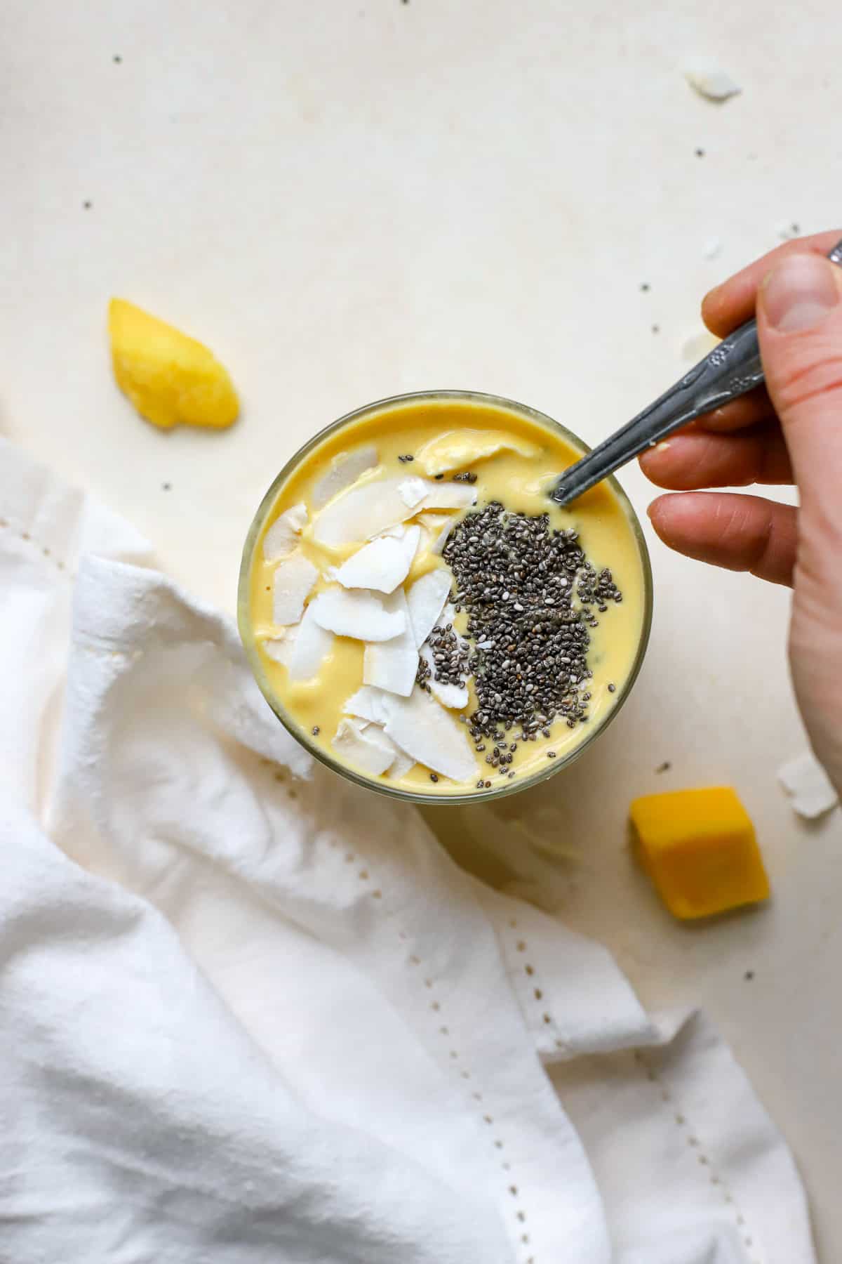 Mango turmeric ginger smoothie topped with coconut chips and chia seeds and hand dipping spoon into smoothie, on beige surface with mango chunks, coconut chips, and white linen.