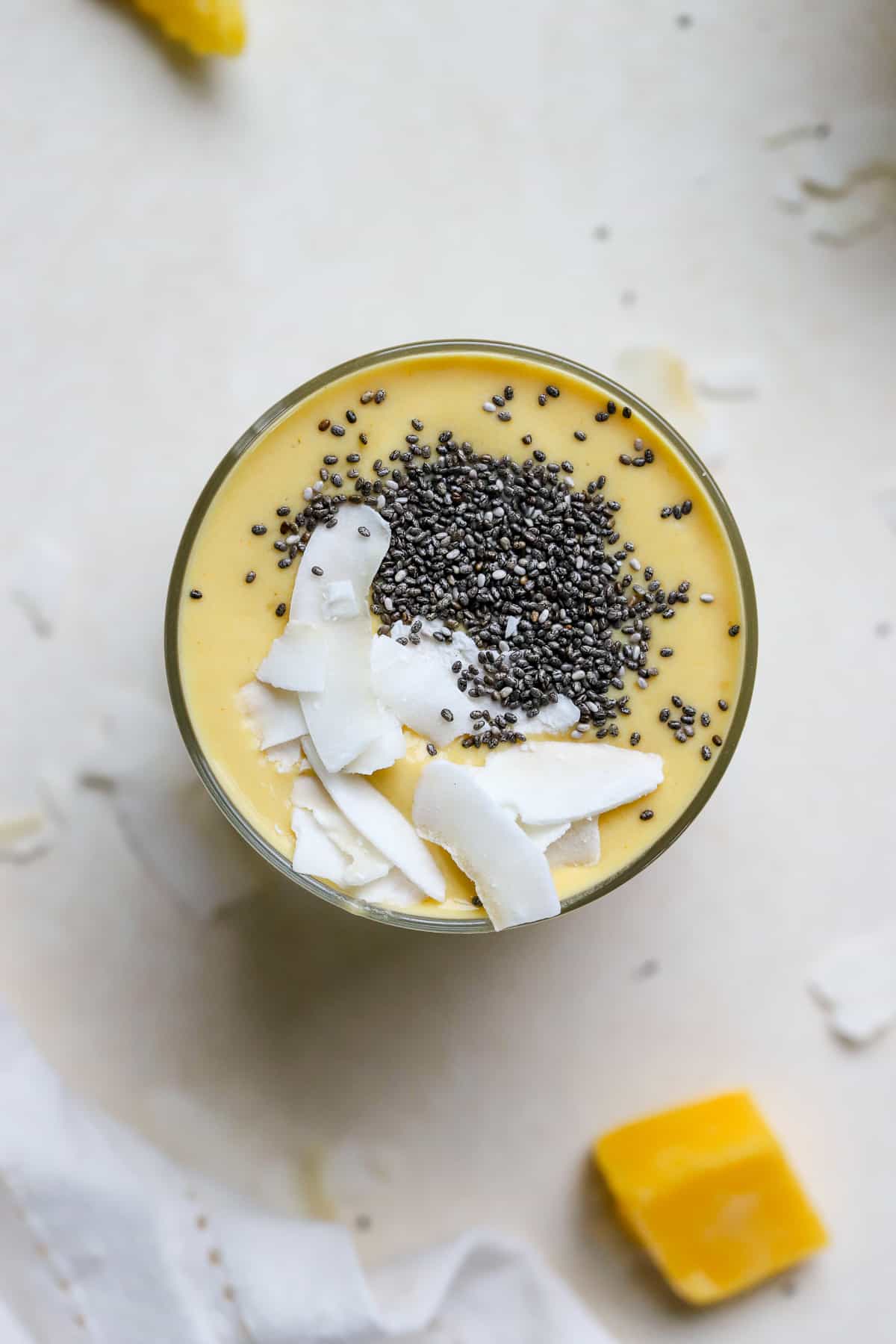 Mango turmeric ginger smoothie topped with coconut chips and chia seeds, on beige surface with mango chunks, coconut chips, and white linen.