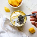 Mango turmeric ginger smoothie topped with coconut chips and chia seeds and hand dipping spoon into smoothie, on beige surface with mango chunks, coconut chips, and white linen.