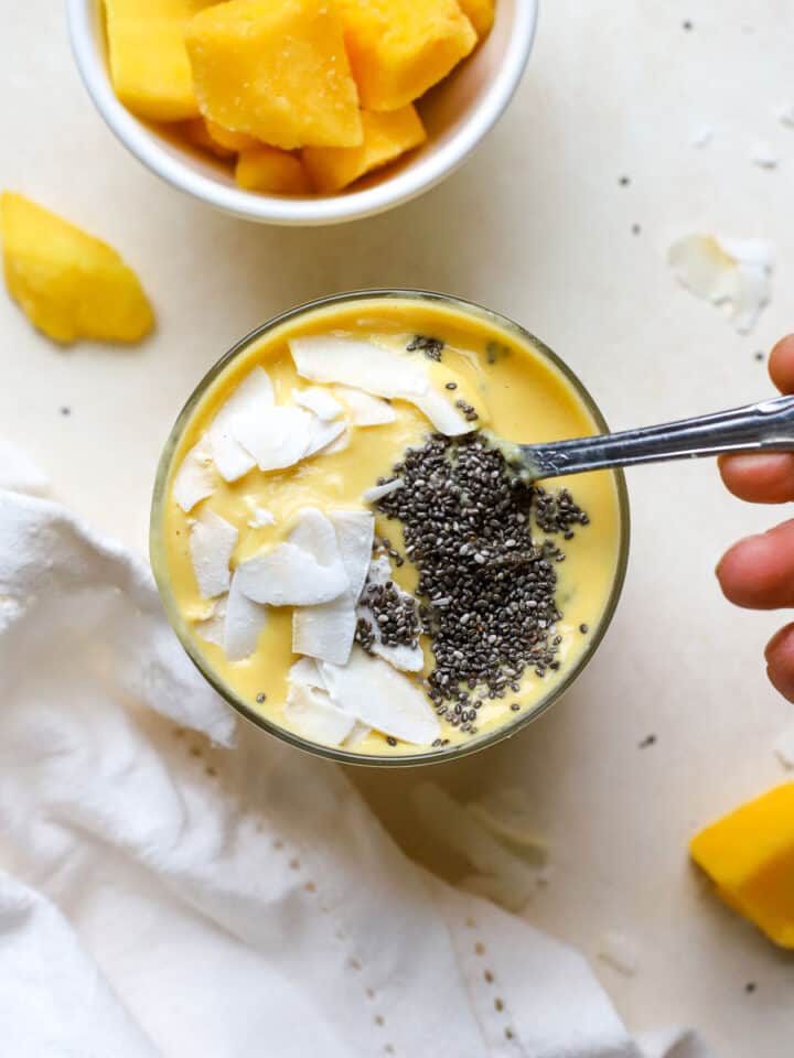Mango turmeric ginger smoothie topped with coconut chips and chia seeds and hand dipping spoon into smoothie, on beige surface with mango chunks, coconut chips, and white linen.