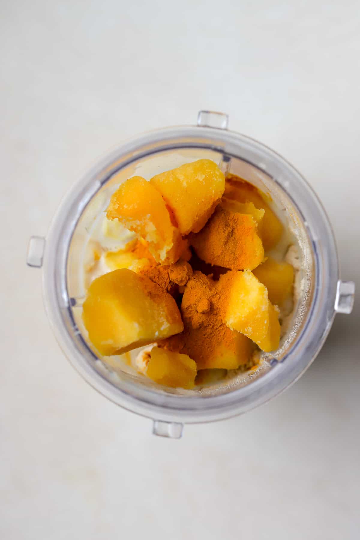 Mango, milk, protein powder, turmeric, and ginger in blender on beige surface.