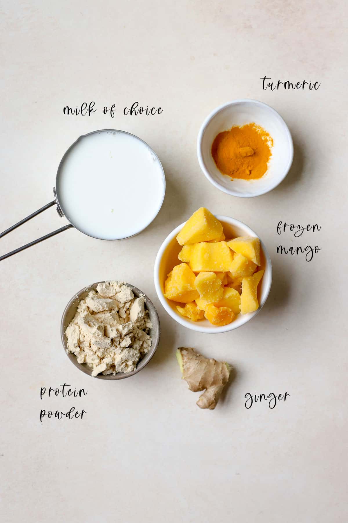 Ingredients for mango smoothies, including milk, frozen mango, ginger root, ground turmeric, and plant-based protein powder, on beige surface.