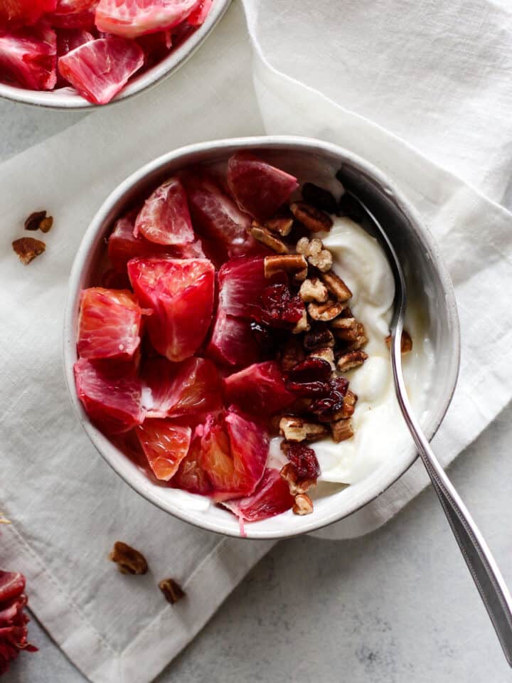 A bowl filled with yogurt and segmented blood oranges piled on the left, pecans and cranberries lined up down the middle, and spoon scooped in on the right. The bowl of yogurt sits on a white linen on a blue and white surface, with a few blood orange segments and pecan crumbs on the side.