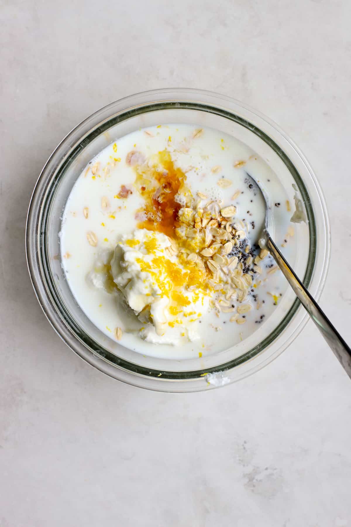 All overnight oats ingredients added to clear glass bowl, not fully mixed yet, with spoon resting in bowl. The bowl sits on a gray and white surface.