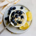 Blueberry lemon overnight oats in a small white bowl topped with juicy blueberries and flecks of lemon zest, and a spoon resting in the oats. The bowl sits on a small blue plate with a few slices of lemon, and a white and yellow windowpane printed towel sits off to the side.