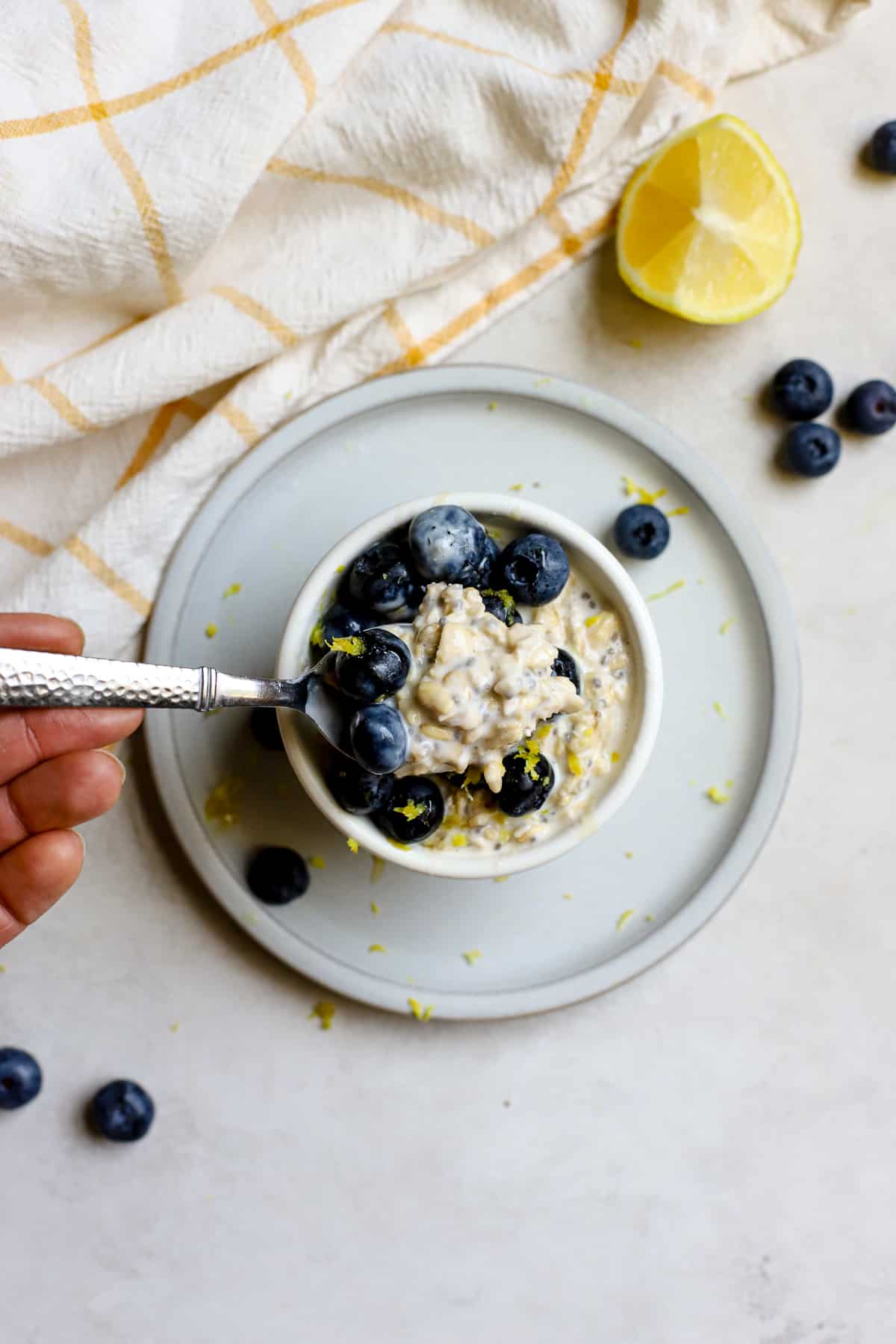 Blueberry lemon overnight oats in small white bowl resting on small blue plate, on gray and white surface. A hand gently starts to scoop a spoonful of oats and blueberries. A few blueberries and a lemon slice sit off to the side with a white and yellow windowpane-printed linen.