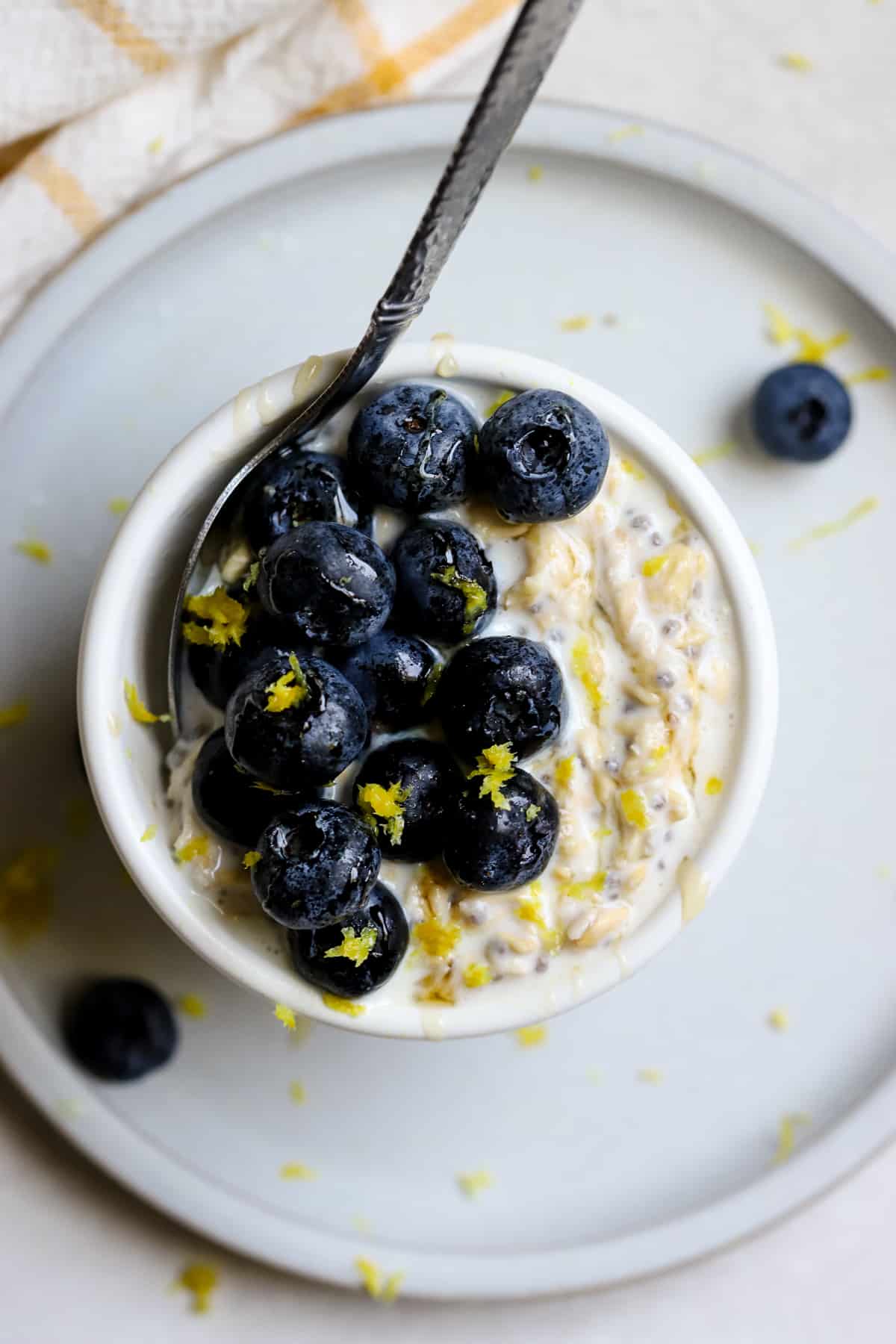 Blueberry lemon overnight oats in small white bowl resting on small blue plate, on gray and white surface. A few blueberries sit off to the side and some pieces of lemon zest is sprinkled around and on top of the oats.