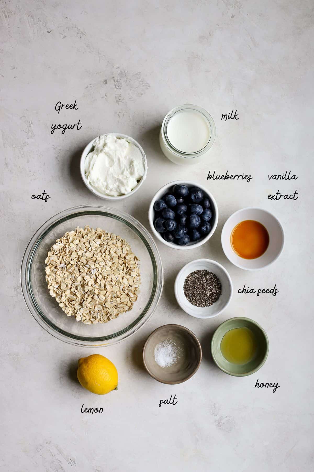 Oats, blueberries, Greek yogurt, milk, vanilla extract, chia seeds, lemon, salt, and honey on gray and white surface.