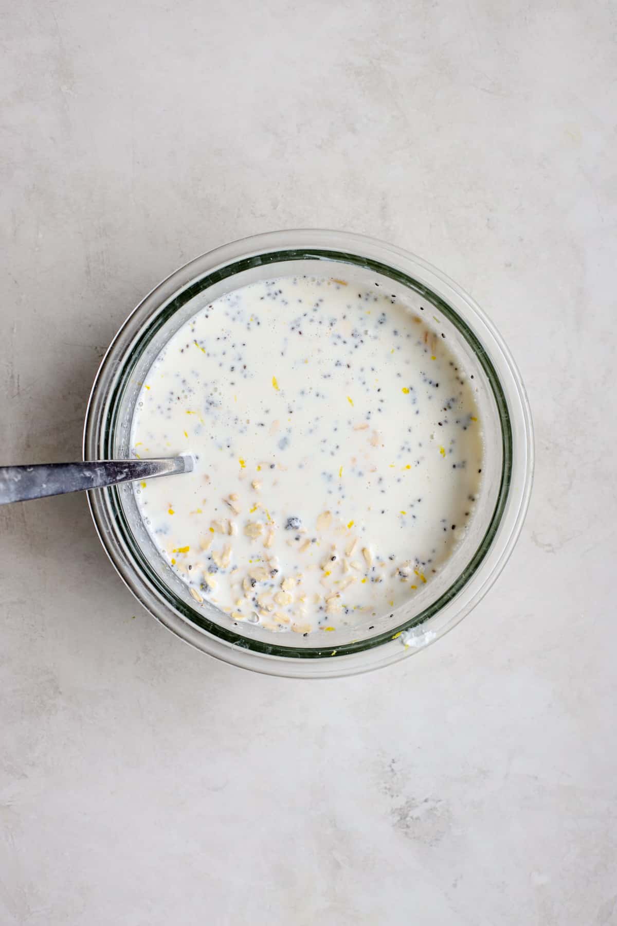 Overnight oats mixture in clear glass bowl with spoon on gray and white surface.