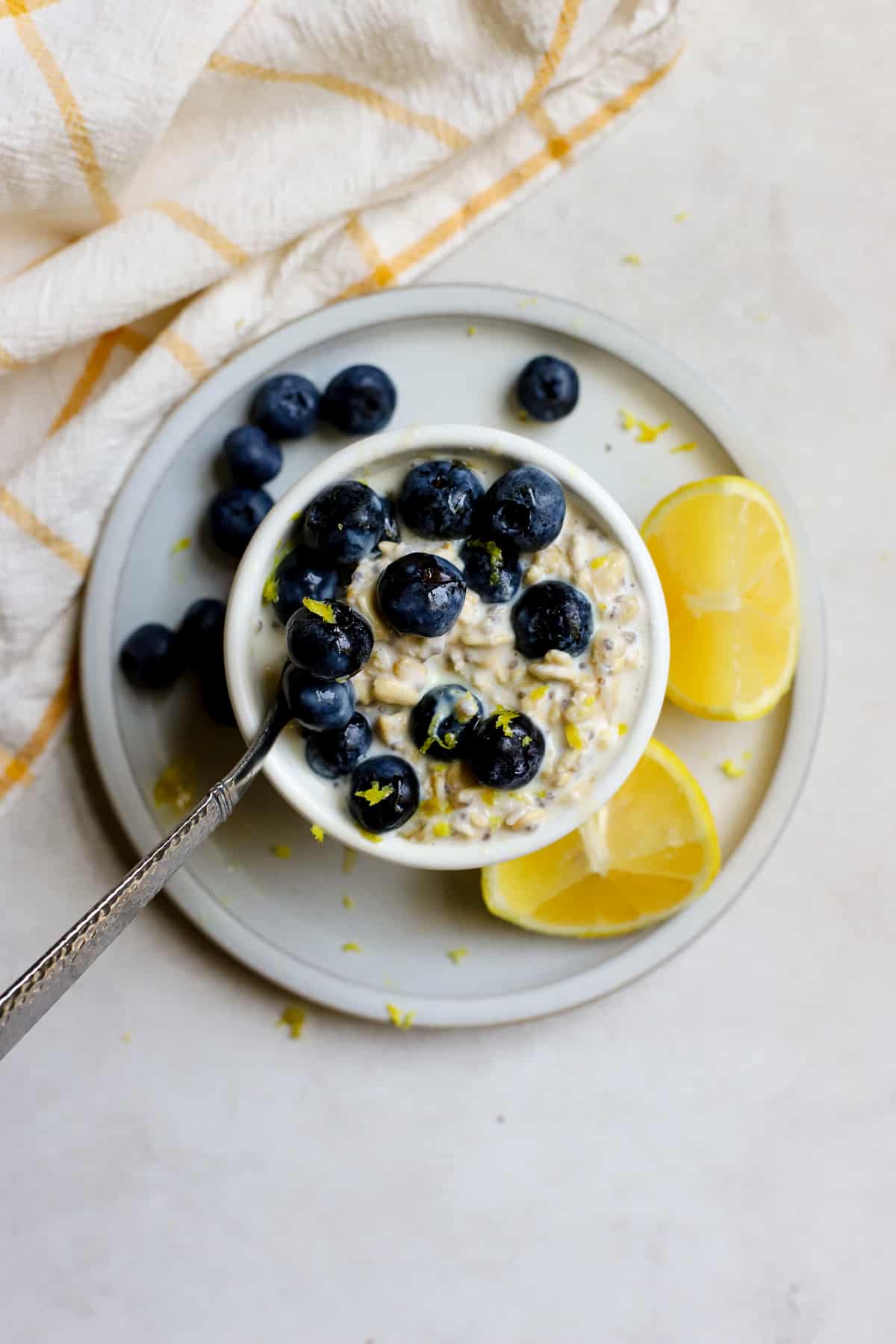 Blueberry lemon overnight oats in a small white bowl topped with juicy blueberries and flecks of lemon zest, and a spoon resting in the oats. 