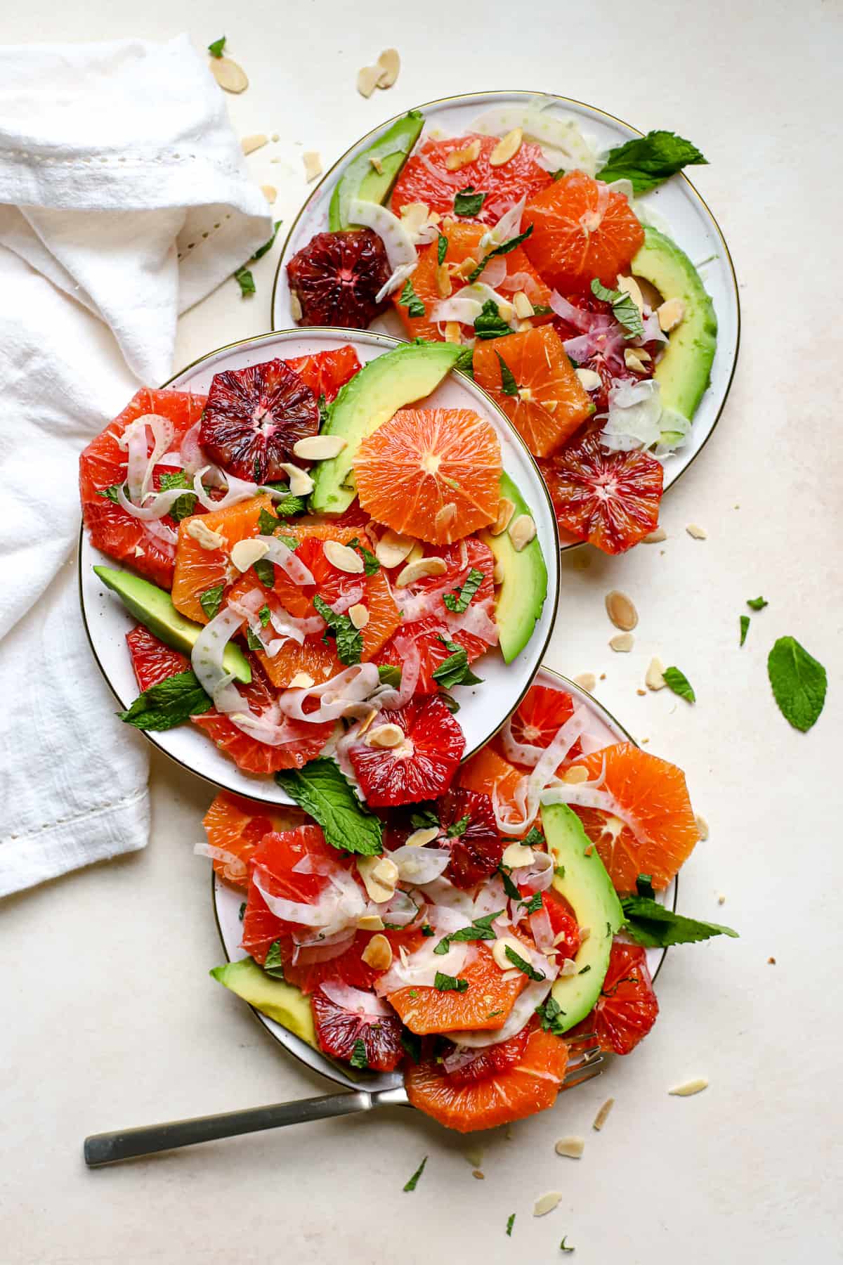 Citrus mint salad with thinly shaved fennel and avocado slices dished up onto three small cream-colored plates with gold edges. The salad is topped with chopped fresh mint leaves and sliced almonds.