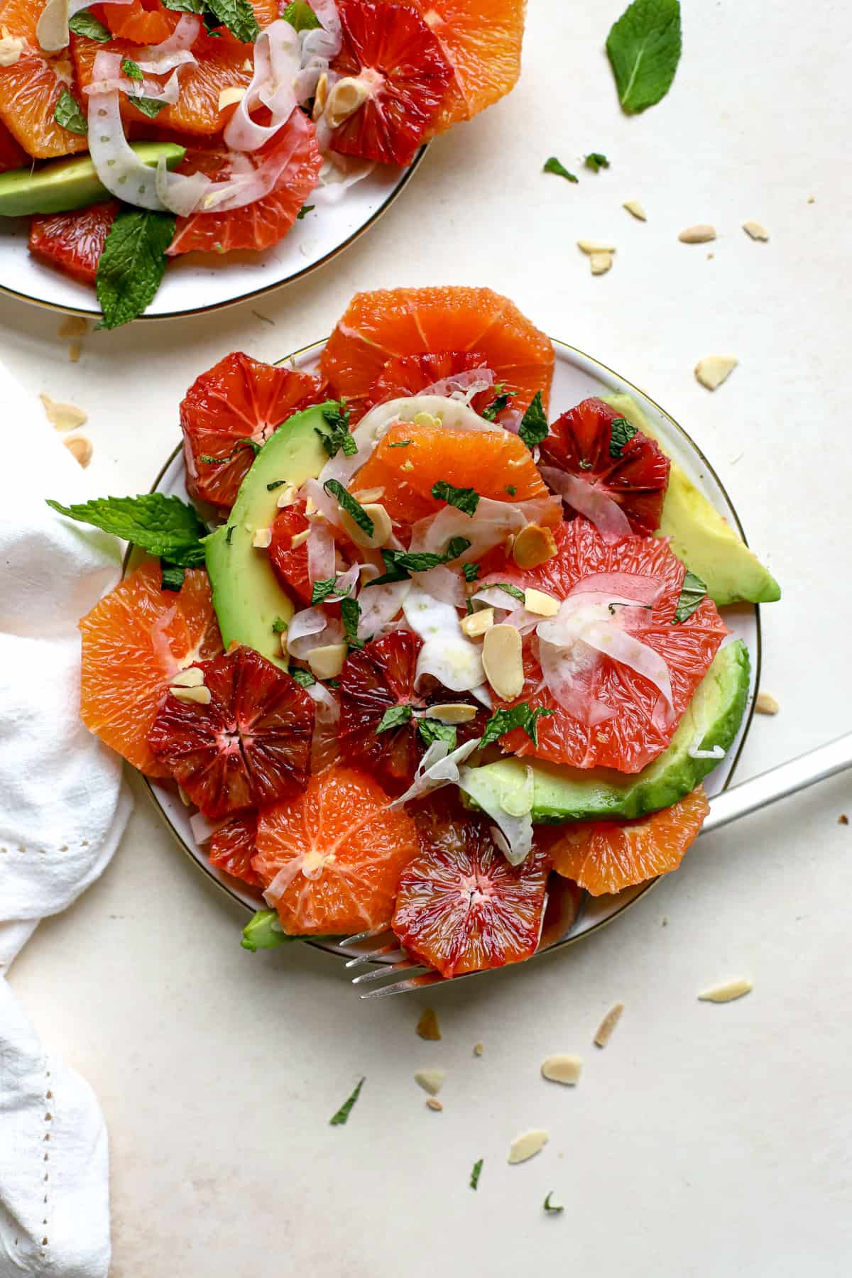 Citrus mint salad with thinly shaved fennel, avocado slices, sliced almonds and chopped mint is served on a cream-colored plate with gold edges. A fork rests on the plate and the plate rests on a beige and cream surface with a few mint leaves and sliced almond crumbs sprinkled around. A white linen rests along the side.