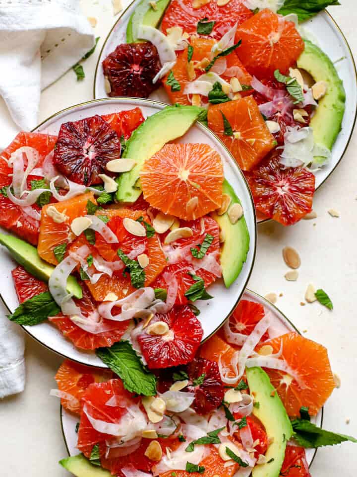 Citrus mint salad with thinly shaved fennel and avocado slices dished up onto three small cream-colored plates with gold edges. The salad is topped with chopped fresh mint leaves and sliced almonds.