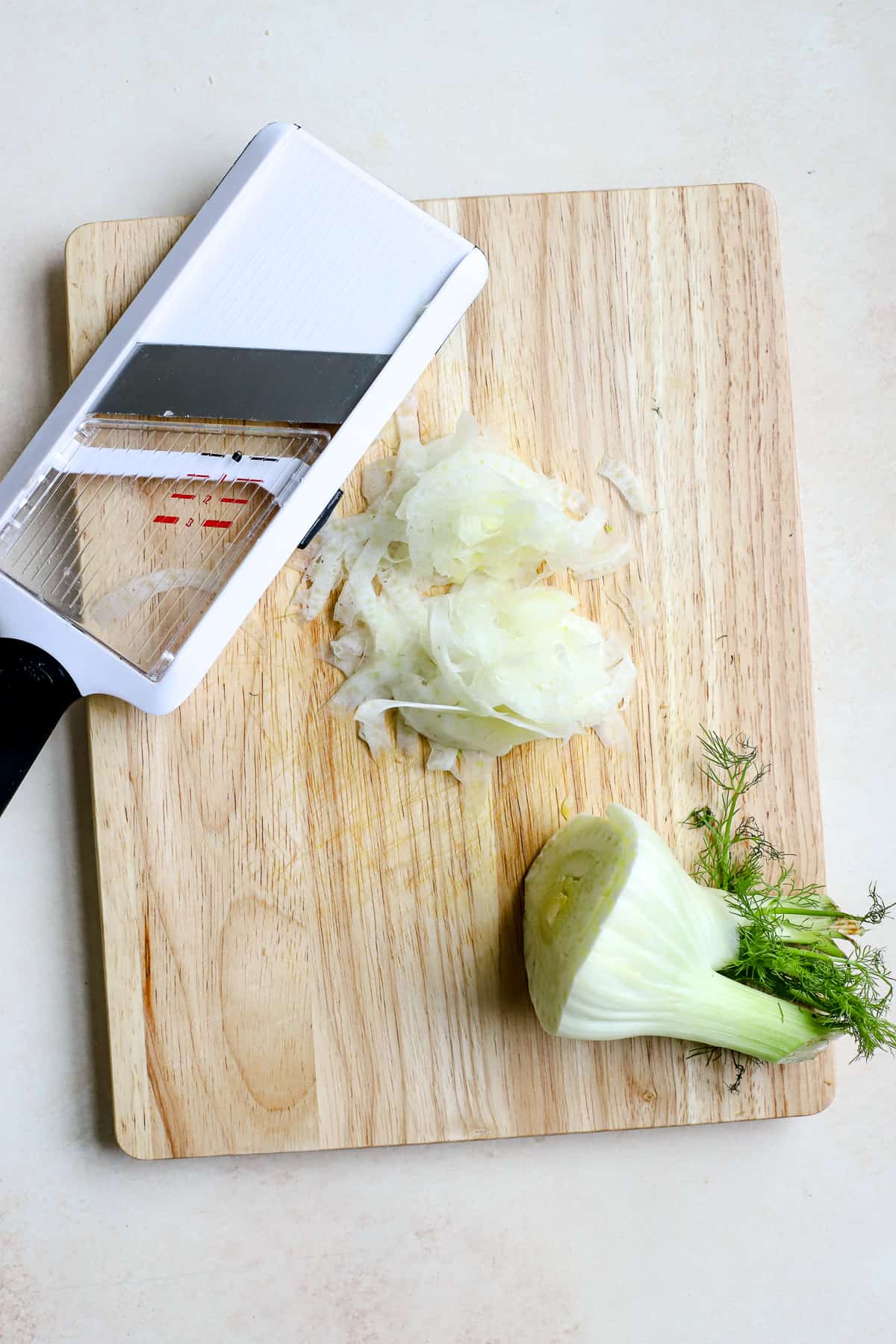 A fennel bulb, mandoline slicer, and fennel shavings all sit on a wooden cutting board, which sits on a beige and cream surface.