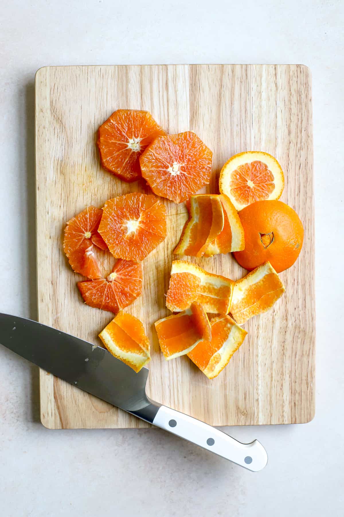 Five round orange slices that are peeled and sliced crosswise through the fruit sit on a wooden cutting board. The peelings and orange ends that were cut off sit off to the side on the same cutting board. The knife used to cut the orange has a white handle and sits on the cutting board by the orange slices.