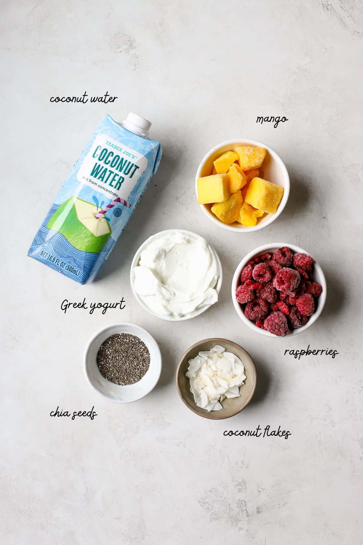 Chia seeds, Greek yogurt, frozen mango, frozen raspberries, and coconut flakes sit on a gray and white surface measured into small bowls. A small carton of coconut water sits next to these ingredients.