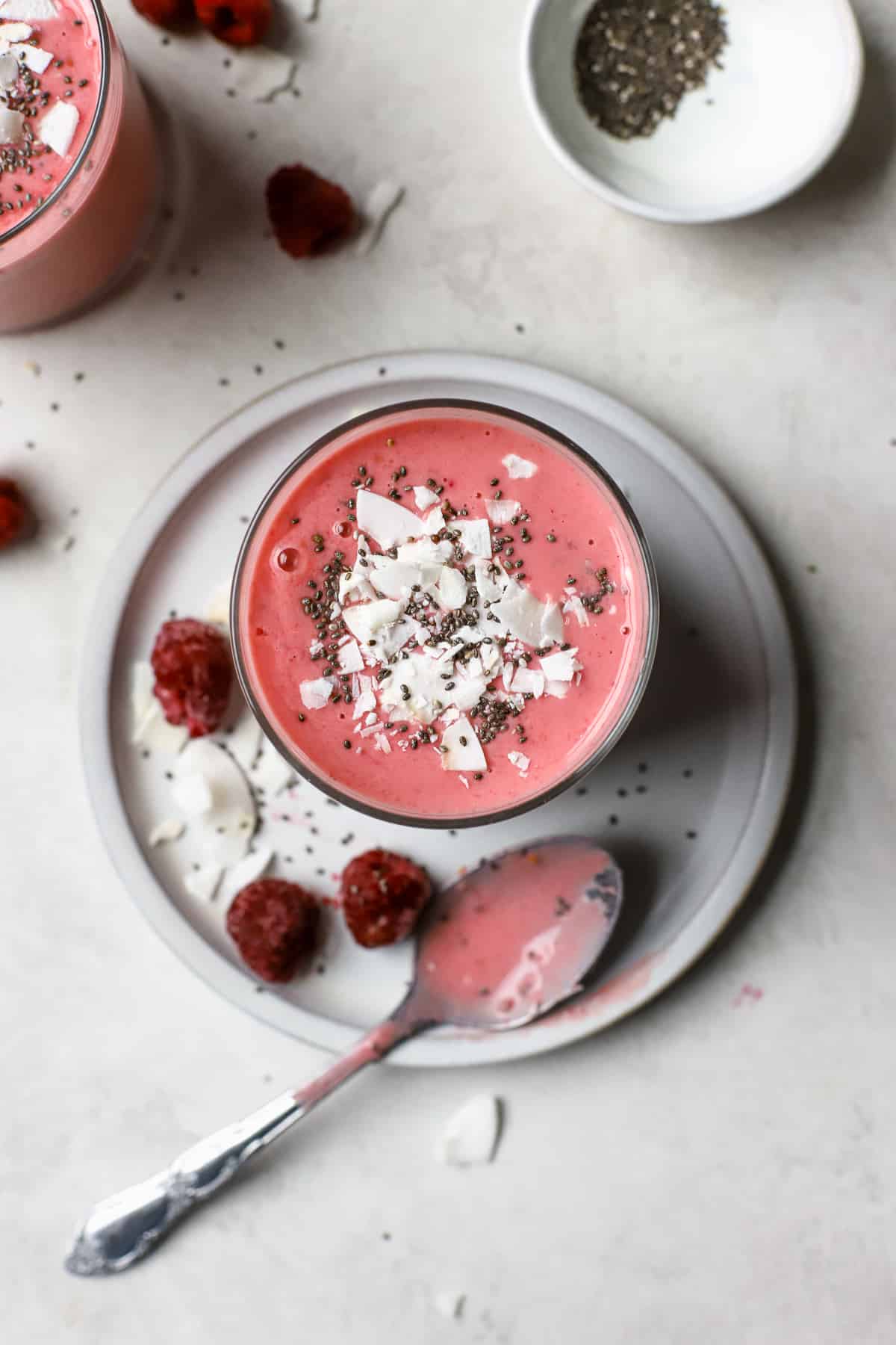 Overhead view of raspberry coconut mango smoothie in small glass with coconut flakes and chia seeds on top. The smoothie sits on a small gray plate with some raspberries, coconut, and chia seeds sprinkled on it, along with a silver spoon that had been dipped in the pink smoothie. Another smoothie sits diagonally to the left and a small white pinch bowl of chia seeds sits diagonally to the right.