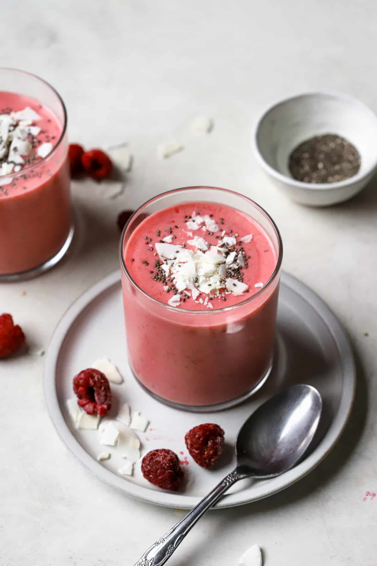 Raspberry coconut mango smoothie topped with coconut flakes and chia seeds, sitting on a small blue plate with a few raspberries and coconut flakes sprinkled around. A silver spoon rests on the plate. Another smoothie sits in the background to the left, while a small white pinch of bowl of chia seeds sits in the background to the right.