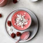 Overhead view of raspberry coconut mango smoothie in small glass with coconut flakes and chia seeds on top. The smoothie sits on a small gray plate with some raspberries, coconut, and chia seeds sprinkled on it, along with a silver spoon that had been dipped in the pink smoothie. Behind this are a smoothie and a small white pinch bowl of chia seeds.