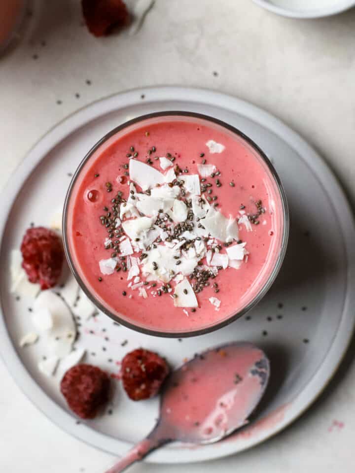 Overhead view of raspberry coconut mango smoothie in small glass with coconut flakes and chia seeds on top. The smoothie sits on a small gray plate with some raspberries, coconut, and chia seeds sprinkled on it, along with a silver spoon that had been dipped in the pink smoothie. Behind this are a smoothie and a small white pinch bowl of chia seeds.