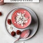 Overhead view of raspberry coconut mango smoothie in small glass with coconut flakes and chia seeds on top. The smoothie sits on a small gray plate with some raspberries, coconut, and chia seeds sprinkled on it, along with a silver spoon that had been dipped in the pink smoothie. Behind this are a smoothie and a small white pinch bowl of chia seeds.