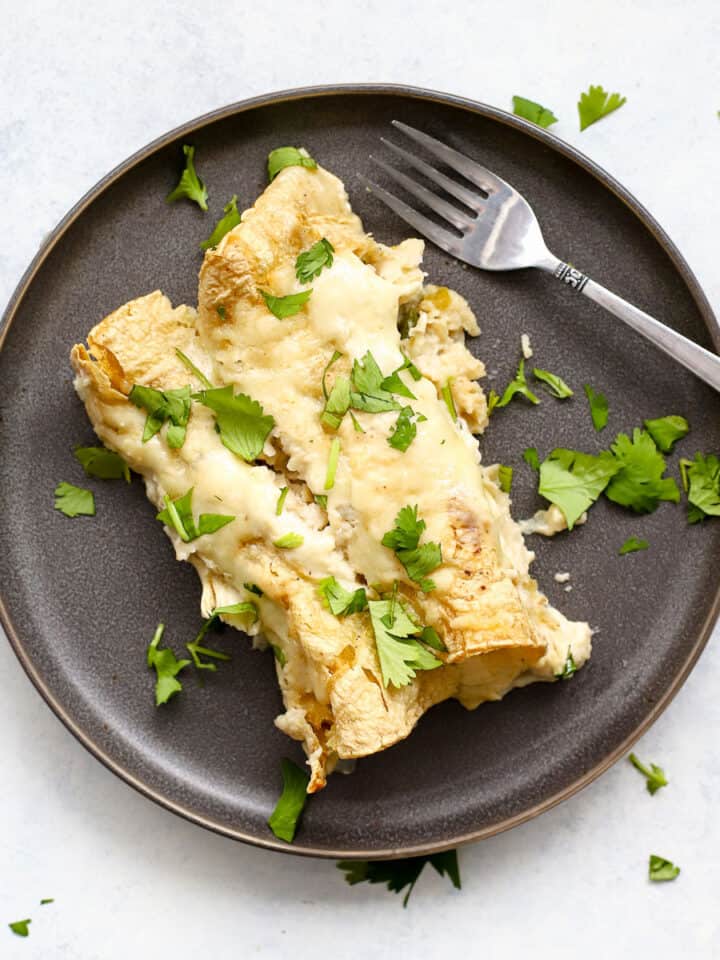 Two healthy white chicken enchiladas on dark gray plate with cilantro sprinkled on side and silver fork resting on plate.