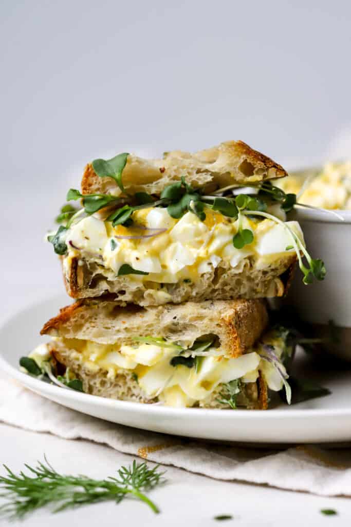 Healthy egg salad with Greek yogurt served as a sandwich on fresh sourdough bread and topped with microgreens. To small sourdough egg salad sandwiches are stacked on top of each other, sitting on a white plate.