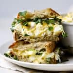 Healthy egg salad with Greek yogurt served as a sandwich on fresh sourdough bread and topped with microgreens. To small sourdough egg salad sandwiches are stacked on top of each other, sitting on a white plate.