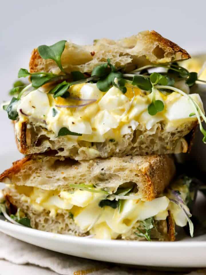 Healthy egg salad with Greek yogurt served as a sandwich on fresh sourdough bread and topped with microgreens. To small sourdough egg salad sandwiches are stacked on top of each other, sitting on a white plate.