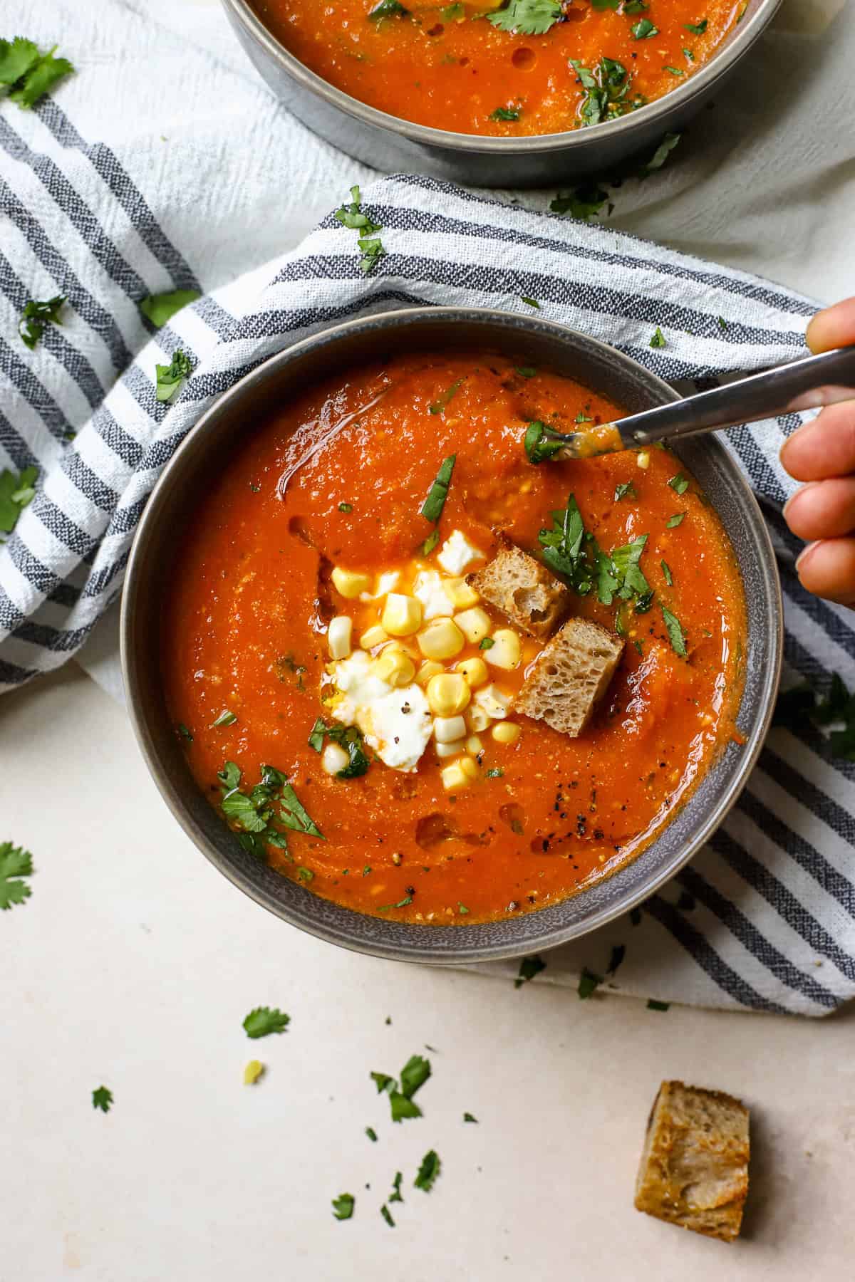 Roasted cherry tomato soup in gray bowl topped with fresh corn, feta, cilantro, pepitas, and croutons.